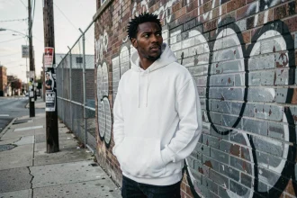 Jeune homme en sweat blanc pensif devant un mur de graffitis dans un quartier urbain défavorisé, illustrant le contexte social du rap de rue