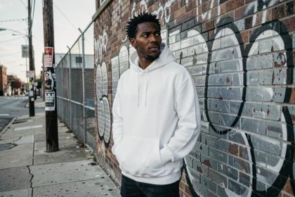 Jeune homme en sweat blanc pensif devant un mur de graffitis dans un quartier urbain défavorisé, illustrant le contexte social du rap de rue